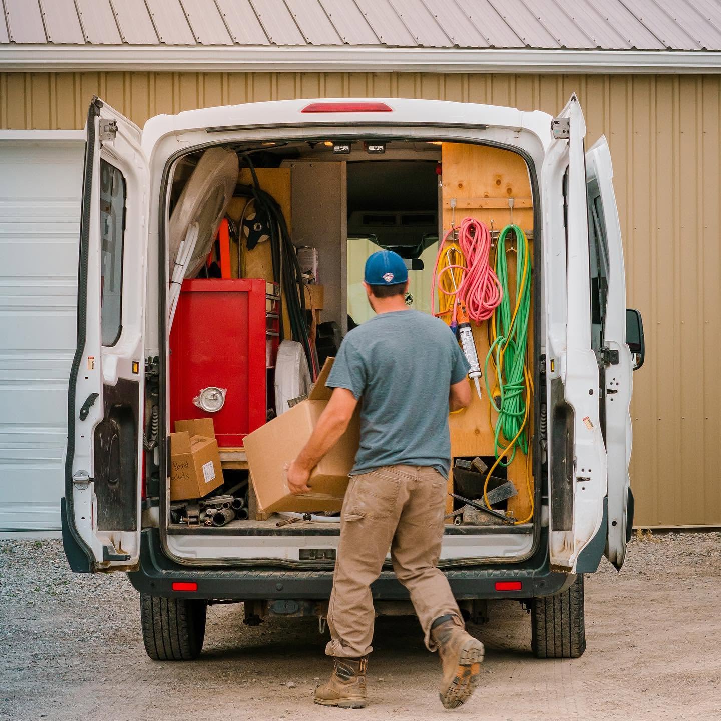 A person in a blue cap and work clothes loads a cardboard box into the back of a white utility van filled with tools, cables, and equipment, parked outside a beige building.