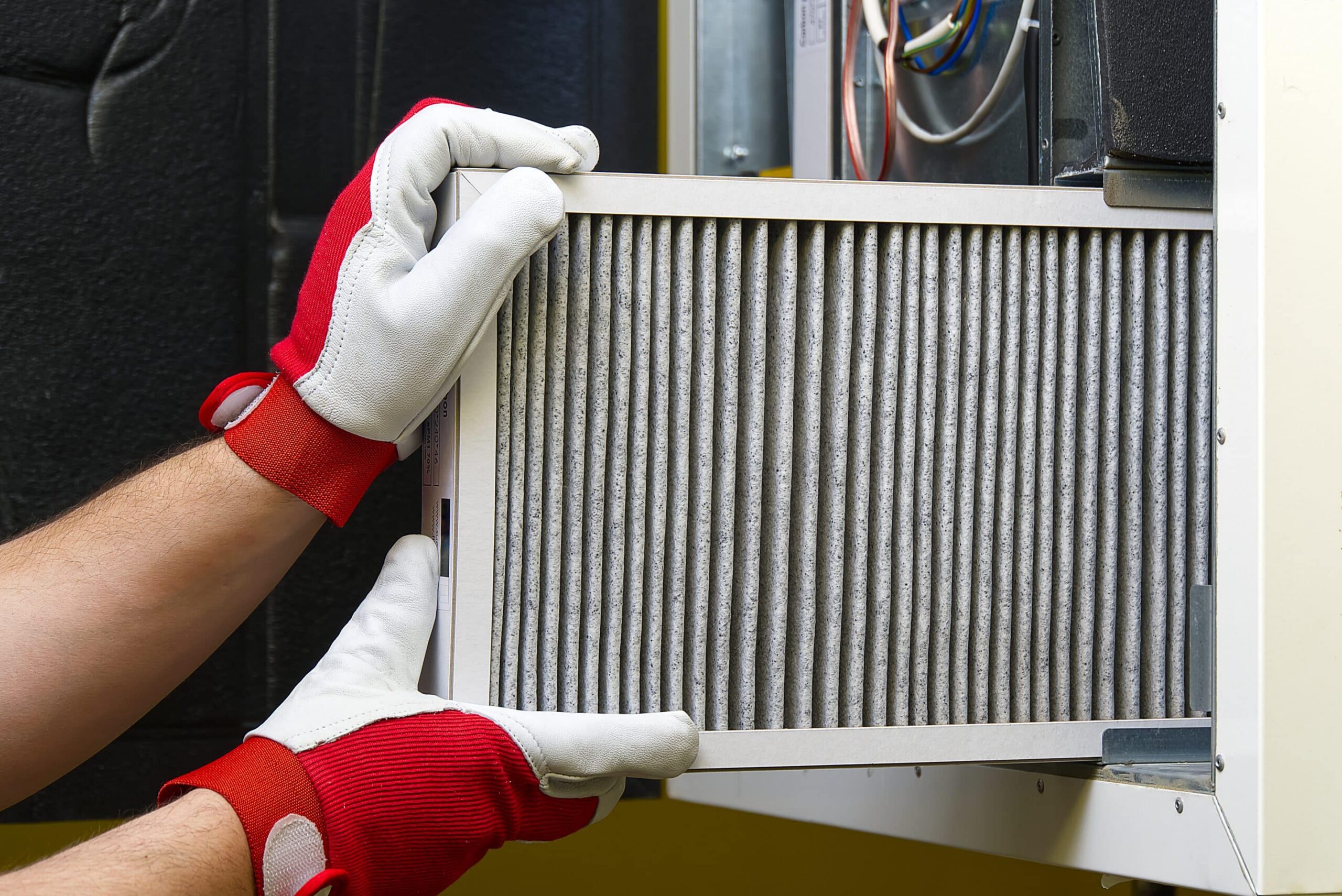 A person wearing red and white gloves is changing your AC filter, inserting or removing a pleated air filter from an HVAC system. The unit's wiring and components are visible in the background.