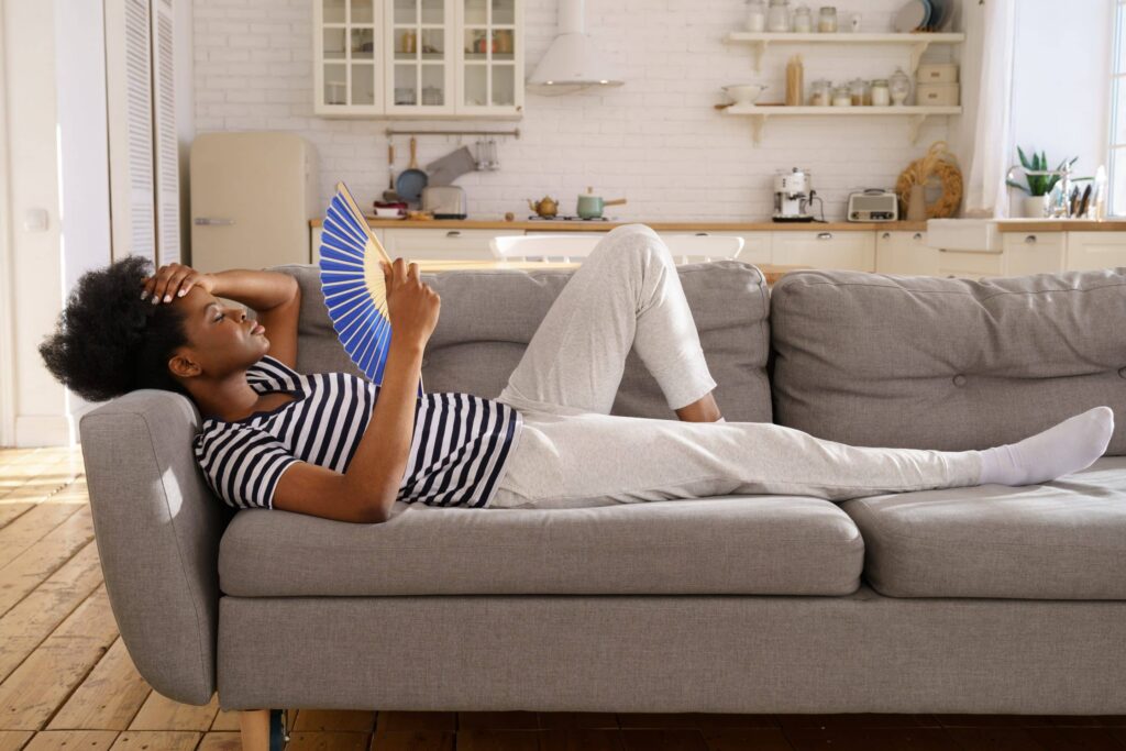 A woman lies on a gray sofa in a bright kitchen, wearing a striped shirt and light pants, holding her head with one hand and fanning herself with a blue fan, looking hot or tired because the AC is blowing warm air.