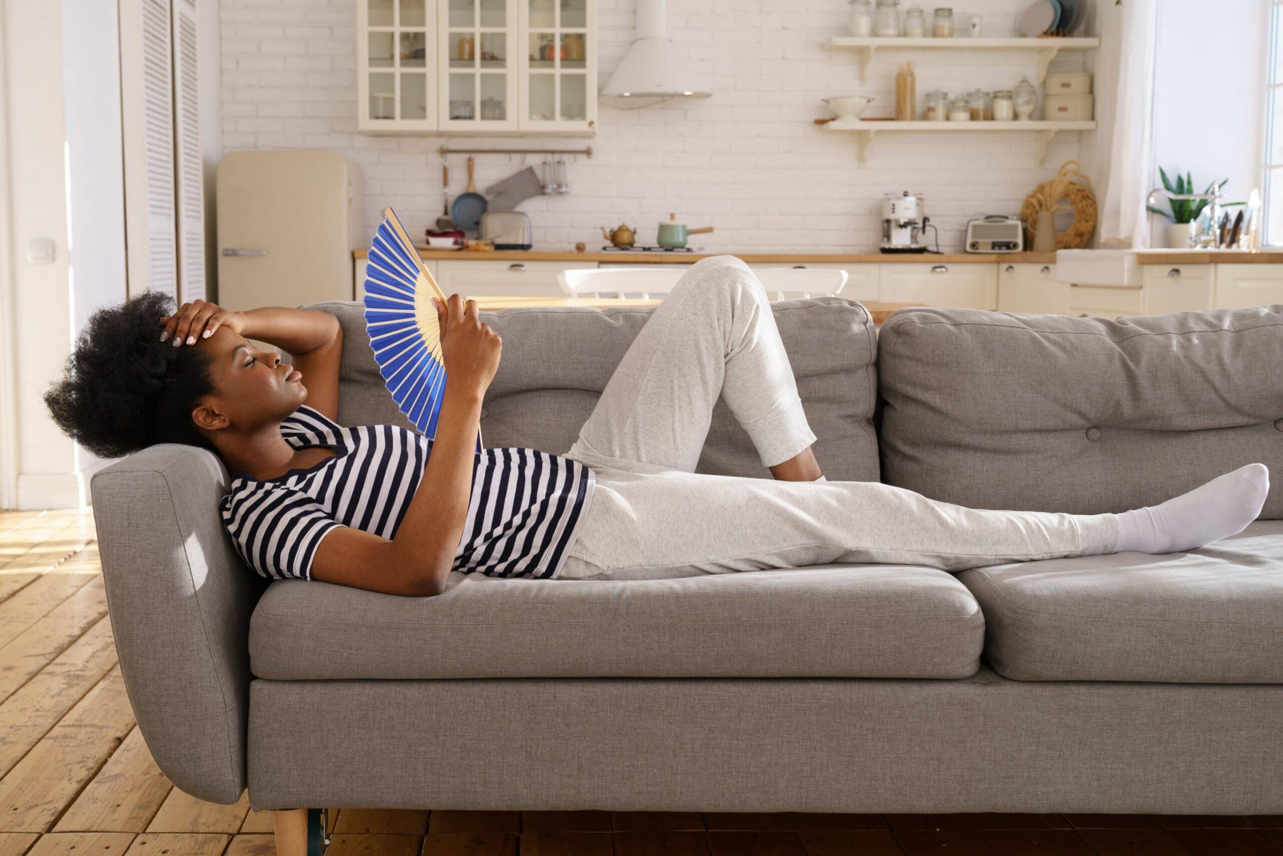 A woman lies on a gray sofa in a bright kitchen, wearing a striped shirt and light pants, holding her head with one hand and fanning herself with a blue fan, looking hot or tired because the AC is blowing warm air.