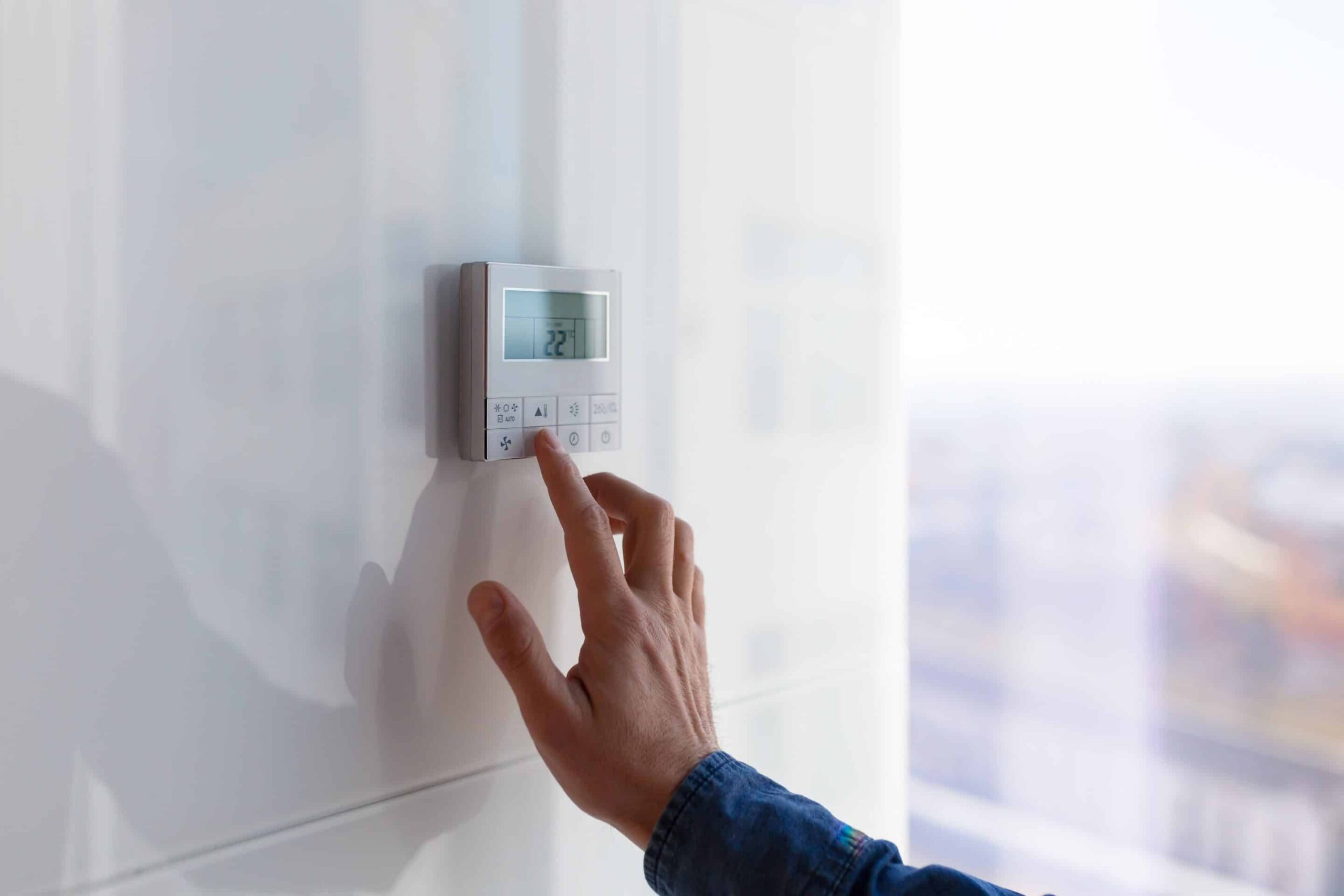 A person’s hand adjusts the temperature on a digital thermostat mounted on a white wall. The display shows “22°C.” Sunlight streams through a window, highlighting how to lower cooling bills this summer with smart temperature control.