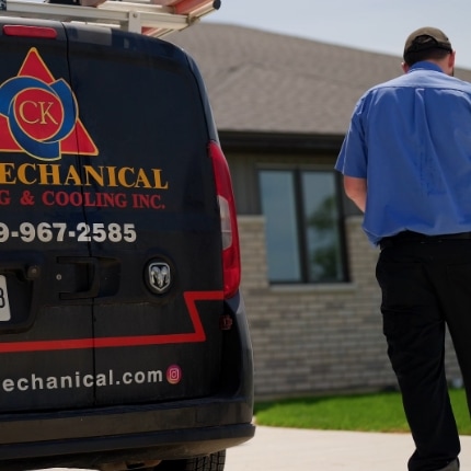 A technician in a blue shirt walks toward a house, next to a black CK Mechanical Heating & Cooling van parked in a driveway on a sunny day.