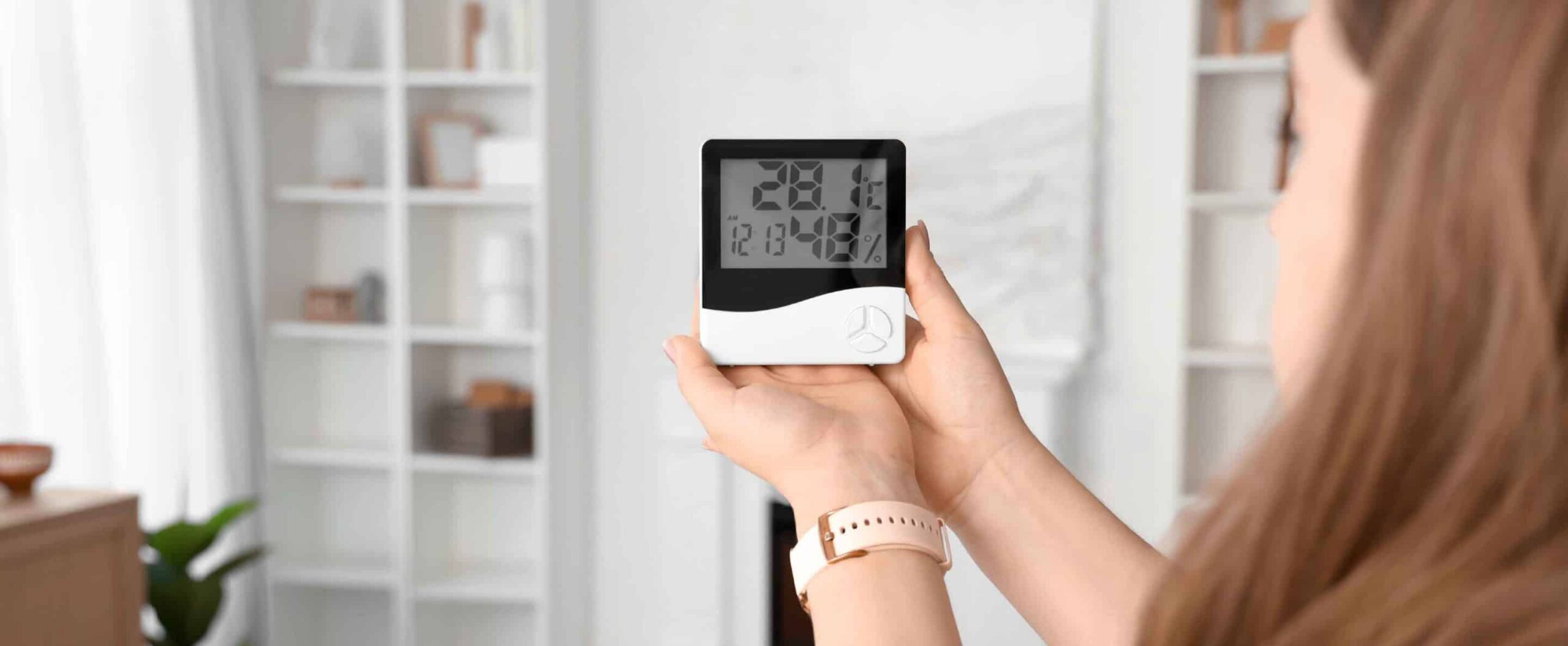 A woman holding a digital thermometer and hygrometer indoors, checking temperature and fall dryness in a modern, bright living room with shelves and decor in the background.