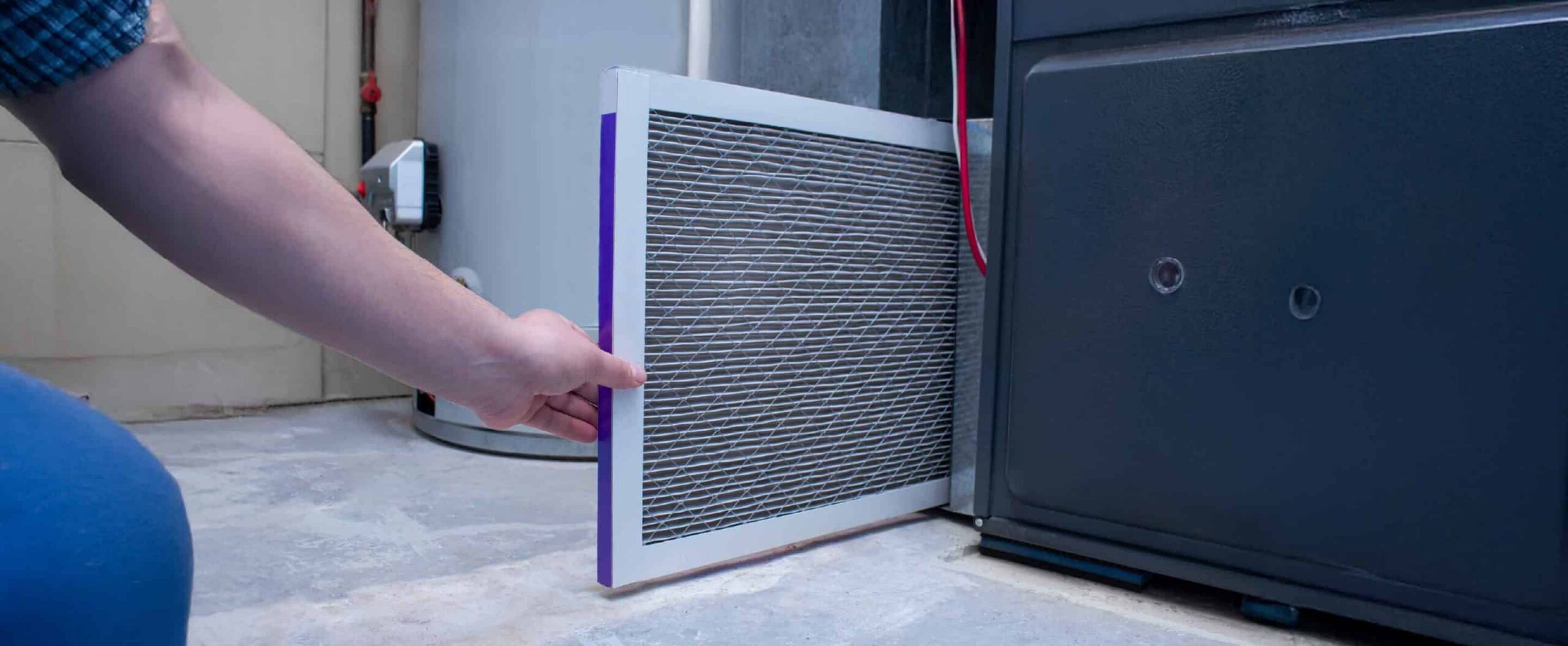 A person’s hand is seen replacing or installing an air filter into a furnace or HVAC unit inside a utility room with a water heater in the background, possibly after their furnace stopped working.