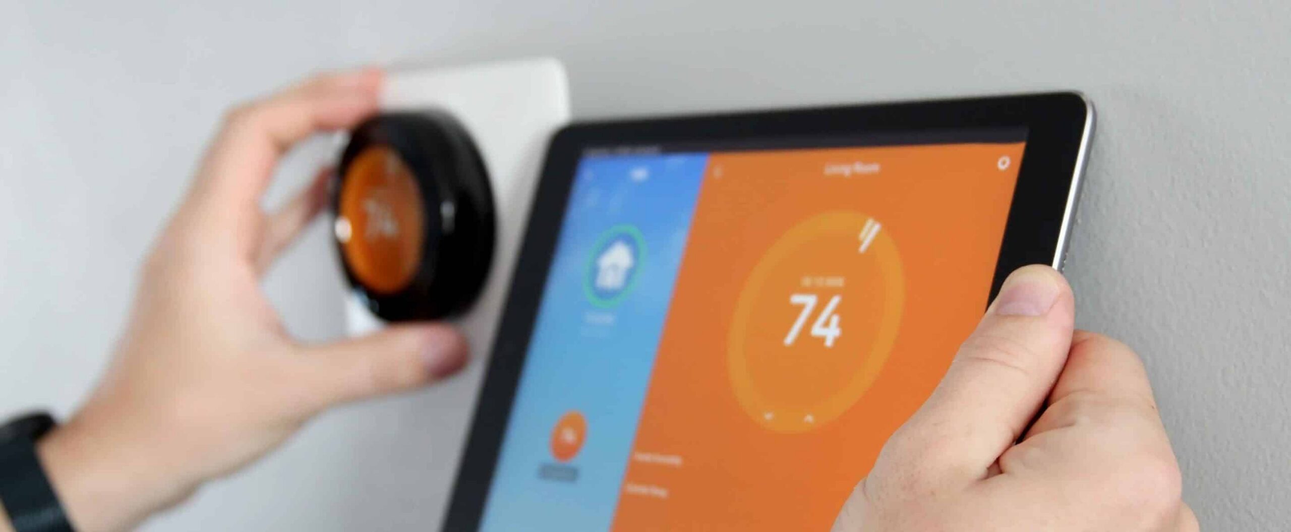A person adjusts a smart thermostat on a wall while holding a tablet displaying the thermostat's 74-degree living room setting, ensuring comfort without leaving the heat on all the time.