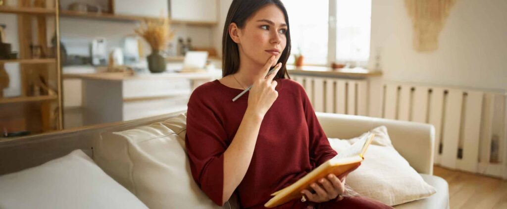 A woman in a red outfit sits on a couch, holding a notebook and pen, thoughtfully preparing her fall HVAC maintenance checklist in a cozy, well-lit living room.