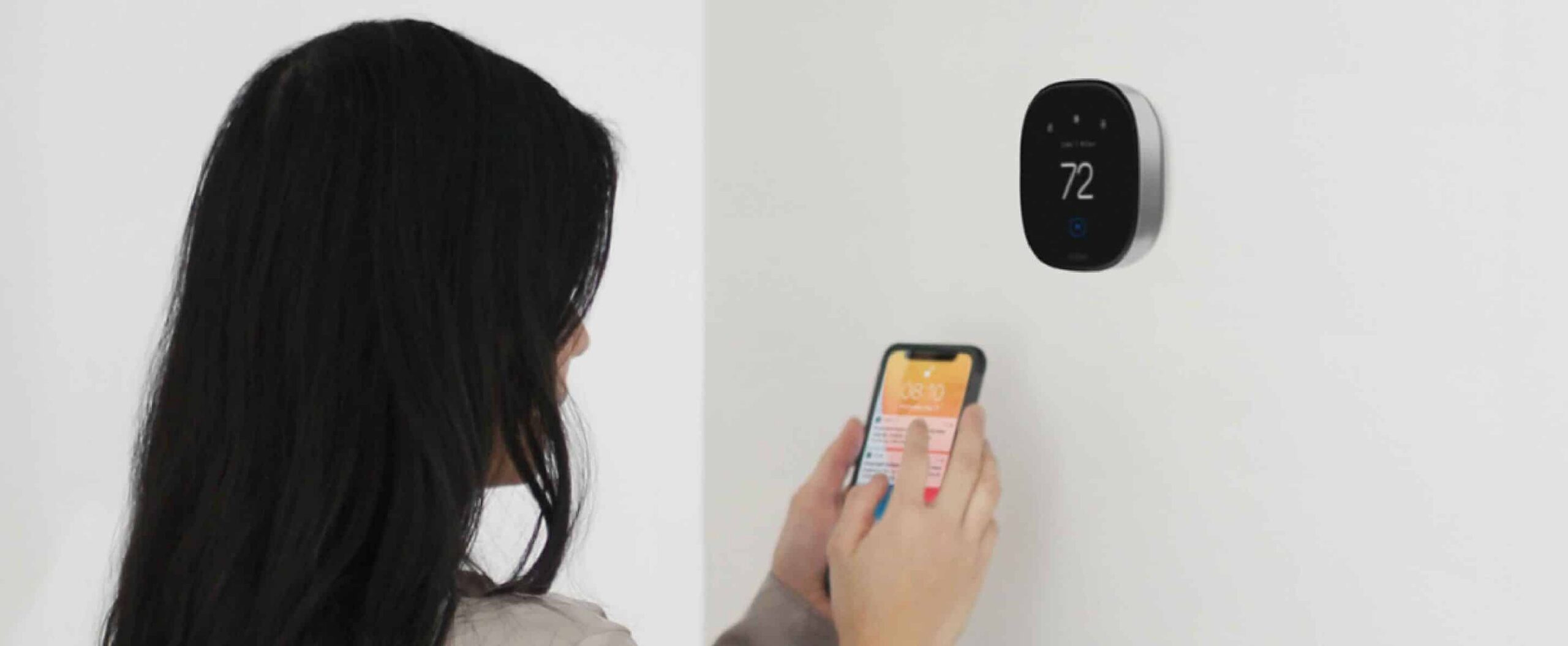 A person with long dark hair uses a smartphone to adjust a smart thermostat mounted on a white wall, displaying 72 degrees and optimized thermostat schedules for winter.