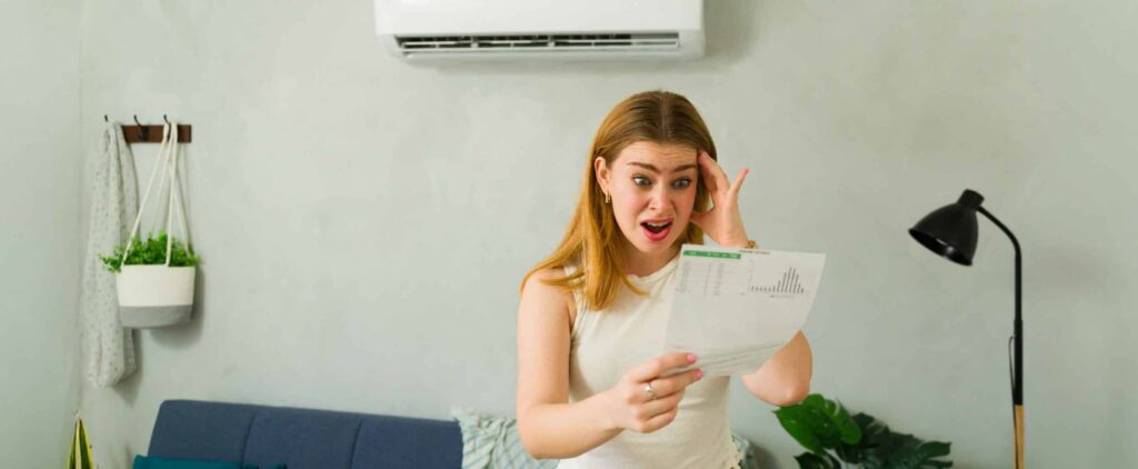 A young woman standing in a living room under an air conditioner looks shocked while reading a bill. She holds one hand to her head, realizing the importance of finding ways to save energy with AC.