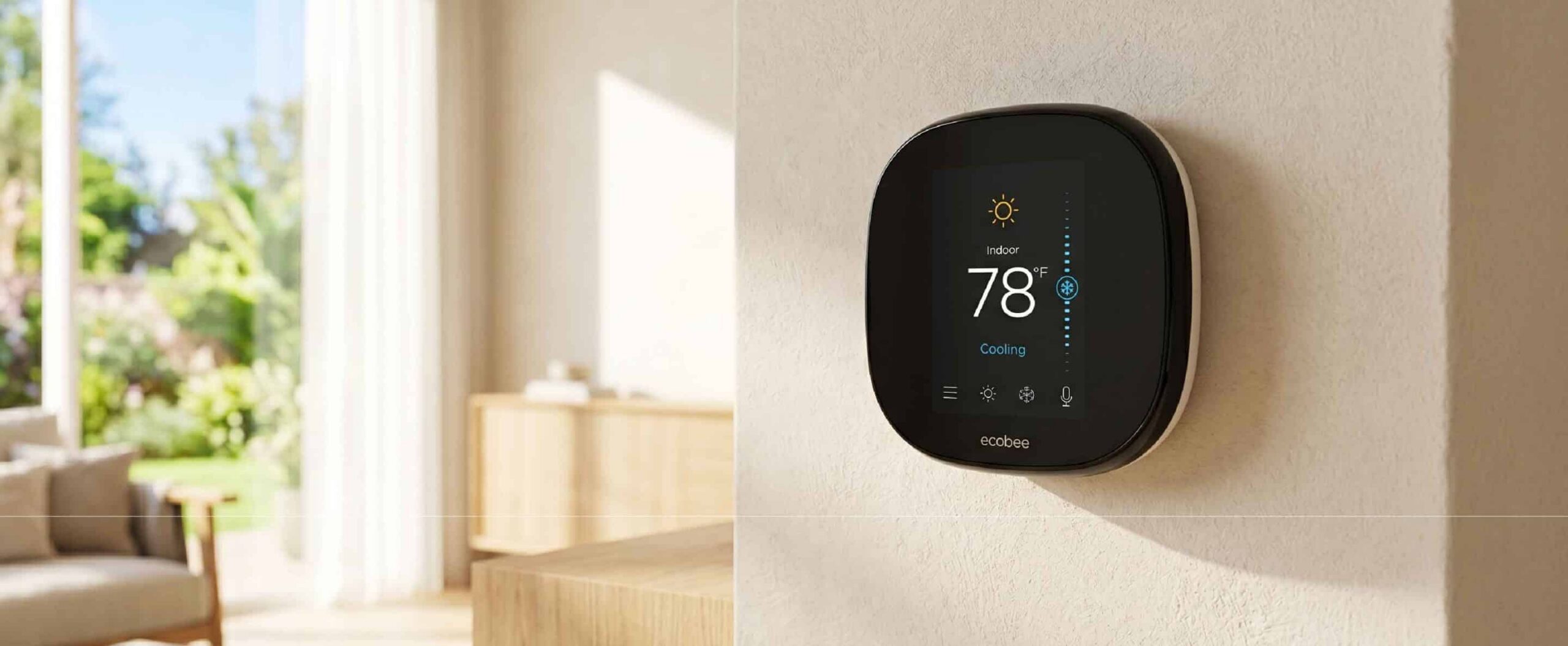 A smart thermostat is mounted on a light-colored wall in a bright, modern living room. The display shows an indoor temperature of 78 degrees—often considered the best AC setting for summer—while cooling mode is active and sunlight streams through large windows.