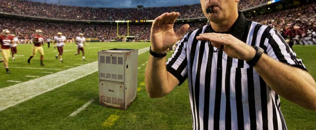 A football referee signals on the field while a large metal industrial furnace, showing furnace failure warning signs, sits on the sideline, with players and a stadium crowd in the background.