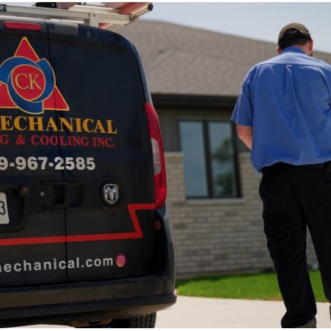 A worker in a blue shirt stands near a black CK Mechanical van parked in front of a modern brick house on a sunny day. The van displays company branding, phone number, and website.