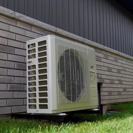 A white outdoor air conditioning unit is installed next to a brick building, slightly elevated above the grass on a metal platform.