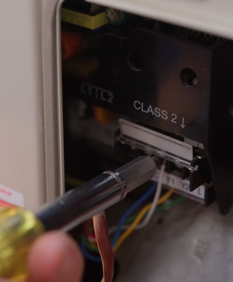 A close-up of a hand holding a screwdriver near an electrical component labeled "CLASS 2," with visible wires and internal parts of a device.