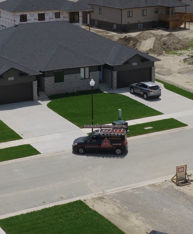A black service van with ladders on its roof is parked in front of a modern, newly built duplex in a suburban neighborhood. Another car is in the driveway and construction materials are visible nearby.