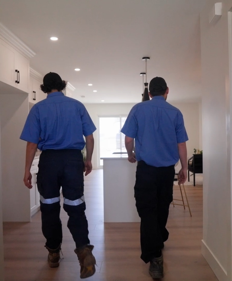 Two people wearing blue shirts, dark pants, and hats walk side by side through a brightly lit, modern kitchen with white cabinets and wooden floors. Their backs are facing the camera.