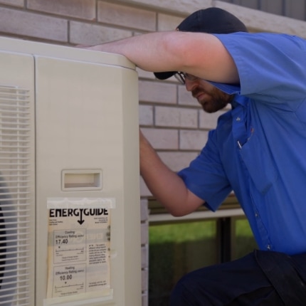 A technician in a blue uniform and cap is inspecting or repairing an outdoor HVAC unit next to a brick wall. An energy guide label is visible on the unit.