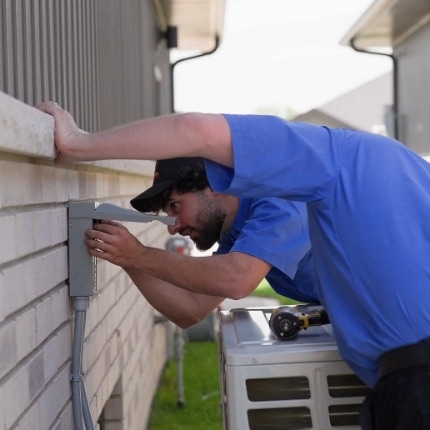 A technician in a blue shirt and cap is working on an electrical box mounted on the exterior wall of a building, with tools and an HVAC unit nearby.