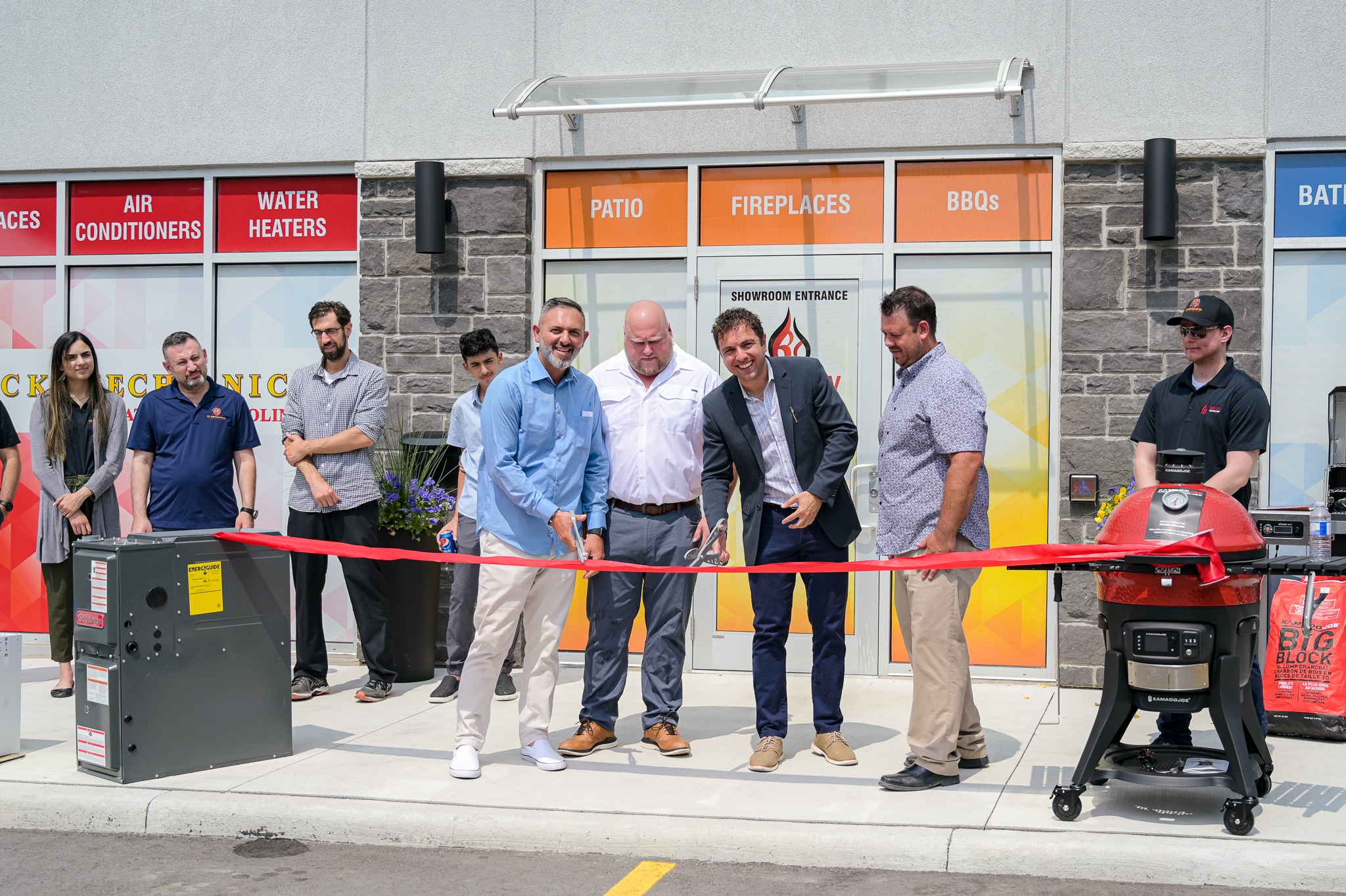 A group of people stand outside a store for a ribbon-cutting ceremony. Two men in the center hold large scissors to cut a red ribbon. Storefront windows behind them display signs for patio, fireplaces, and BBQs.