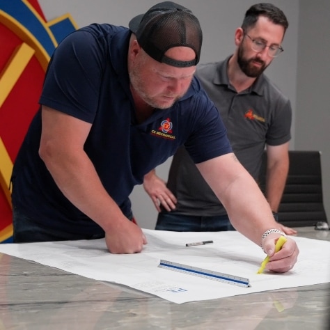 Two men review large architectural plans on a table. The man in front is highlighting the document with a yellow marker, while the other observes. Both wear casual clothes and appear focused on the work.