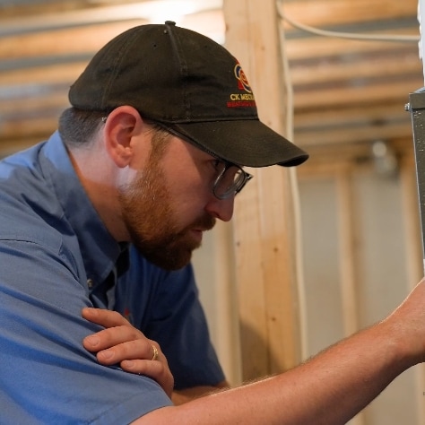 A man wearing glasses, a black cap, and a blue shirt examines an electrical panel in an unfinished room with exposed wooden beams.