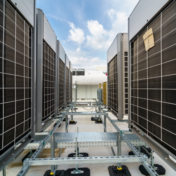Rows of large industrial air conditioning units installed on a rooftop, with metal frames supporting the equipment. The sky is partly cloudy in the background.