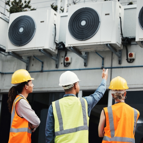 Three workers wearing safety vests and hard hats stand outside, looking up at and discussing a row of outdoor air conditioning units mounted on a building wall.