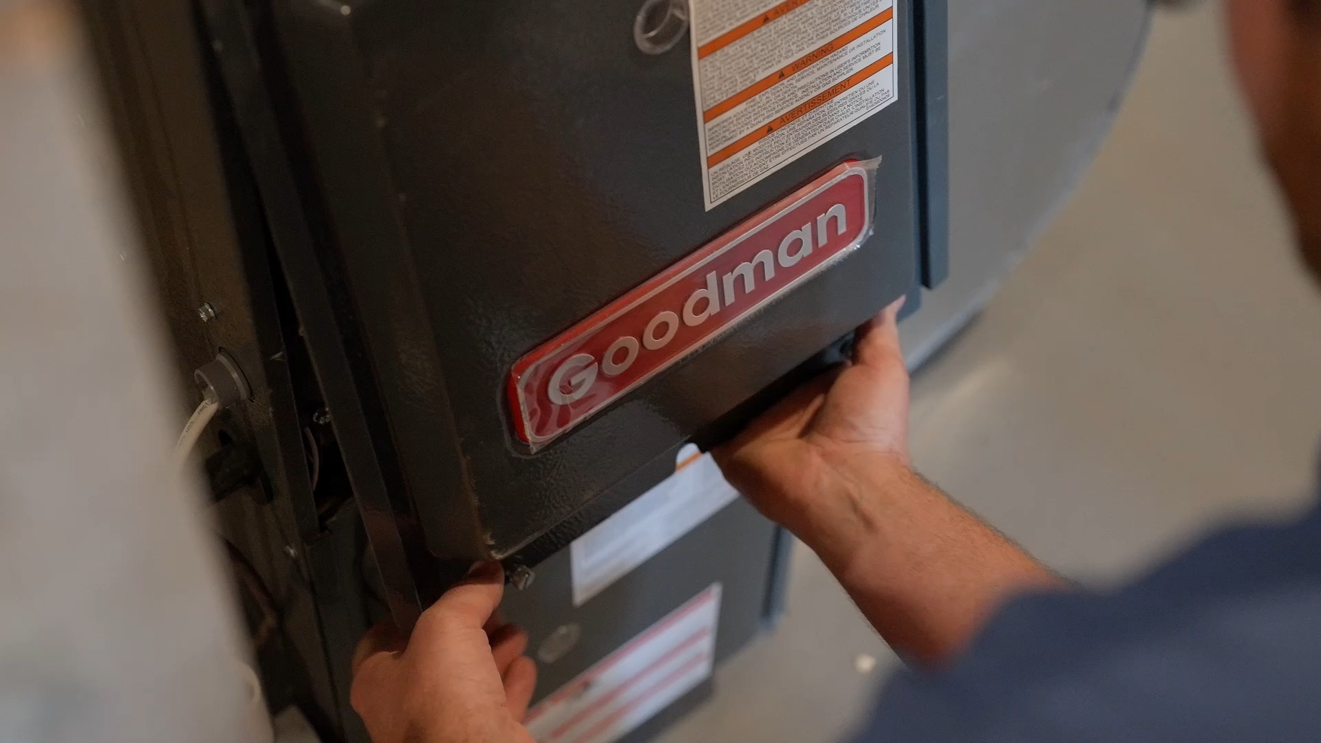 A person is removing the front panel of a Goodman furnace, exposing the unit for maintenance or repair. The furnace has safety and instruction labels visible on its surface.