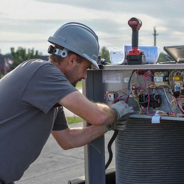 A technician wearing a hard hat and gloves repairs electrical components inside an outdoor HVAC unit, with tools and wires visible.