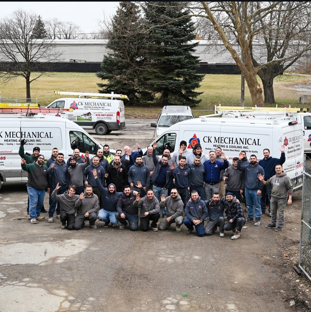 A large group of workers poses and smiles for a photo in front of several CK Mechanical service vans in an outdoor parking lot, with trees and a building in the background.