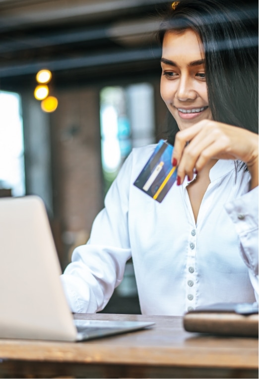 A woman in a white shirt smiles while holding a credit card and using a laptop, suggesting she is shopping or making a payment online in a modern, well-lit setting.