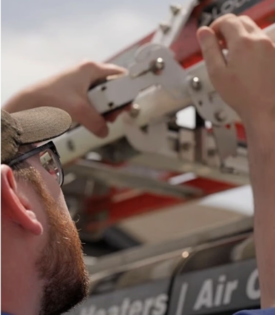 A person wearing glasses adjusts a metal lever on a mechanical structure outdoors, with a partial view of signage and sky in the background.